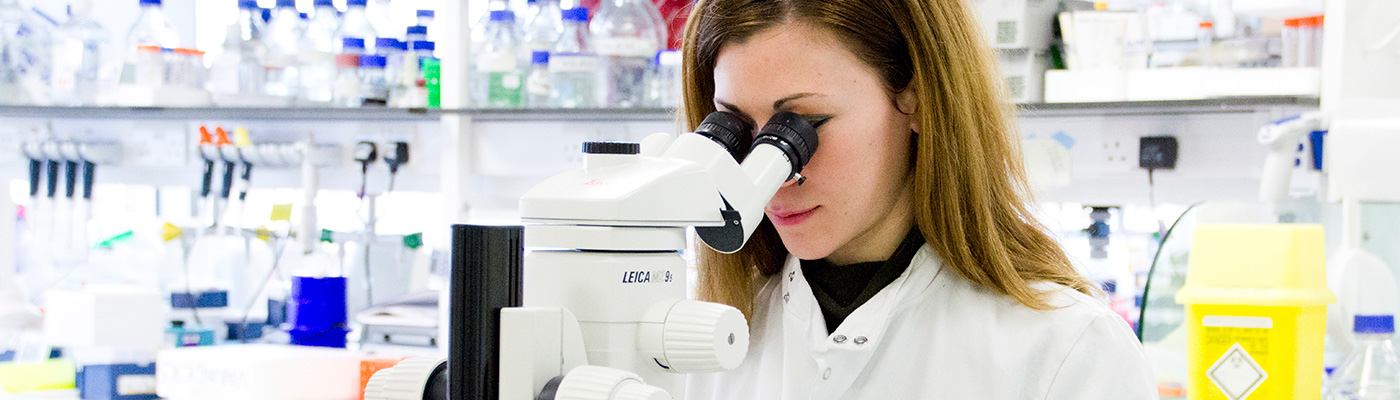 Female researcher in white lab coat peering down microscope