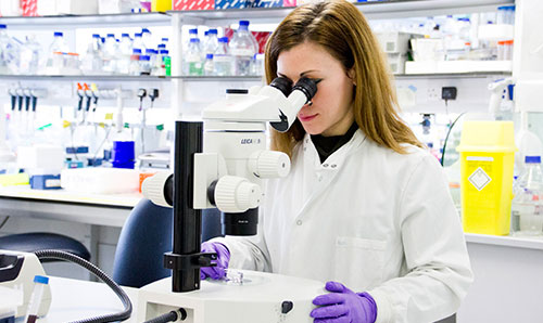Researcher in white lab coat peering down microscope